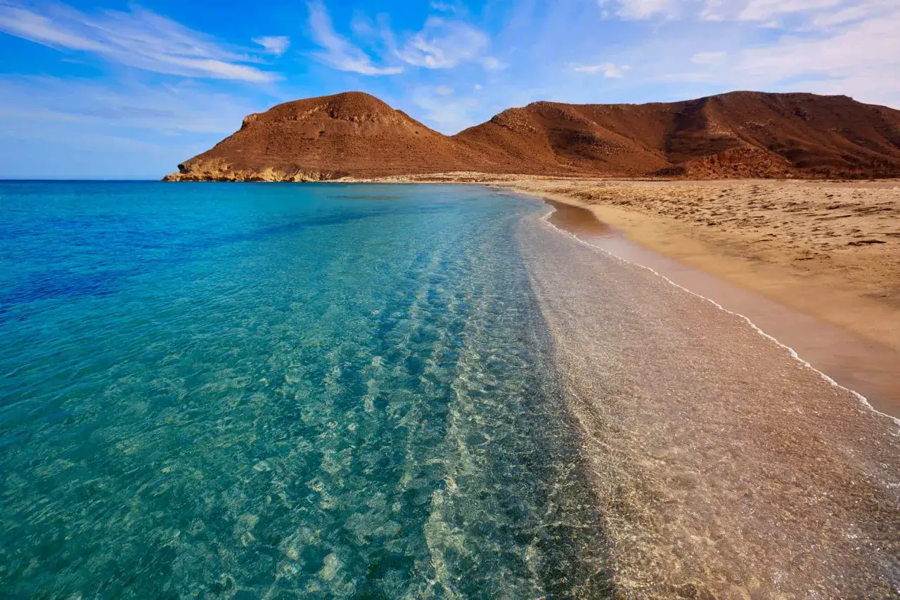 Aguas cristalinas y arena dorada en una de las mejores playas de Almería, con montañas áridas al fondo bajo un cielo azul.