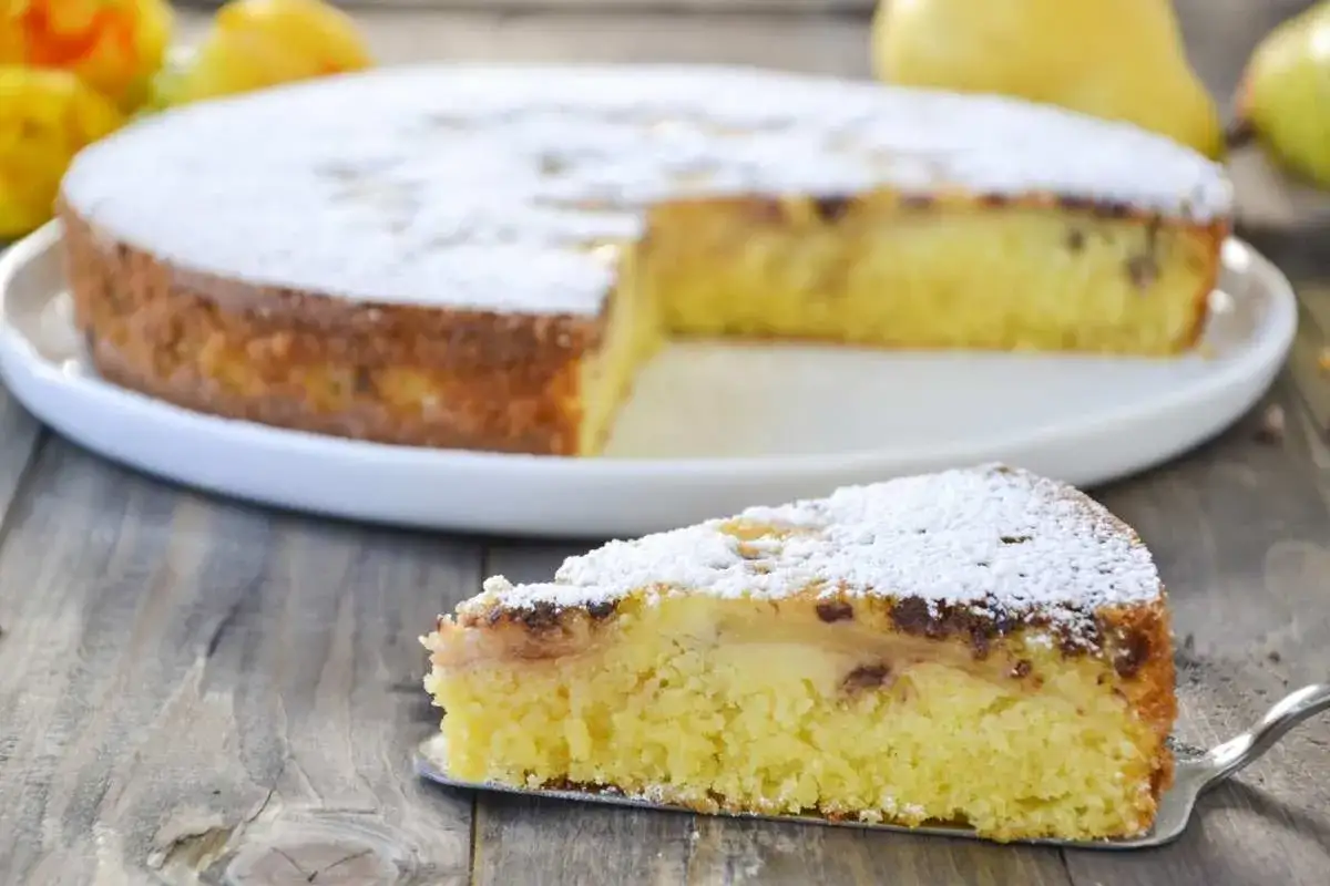 Una fetta di torta pere e cioccolato, cosparsa di zucchero a velo, su un piatto da portata. Sullo sfondo, la torta intera.
