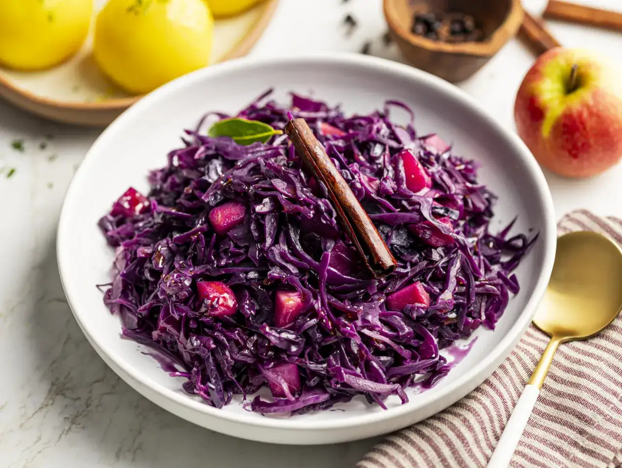 Schmorgerichte mit rotkohl verfeinern: Ein Teller mit gedünstetem Rotkohl, Apfelstücken und Zimtstange, daneben ein Apfel und Gewürze.