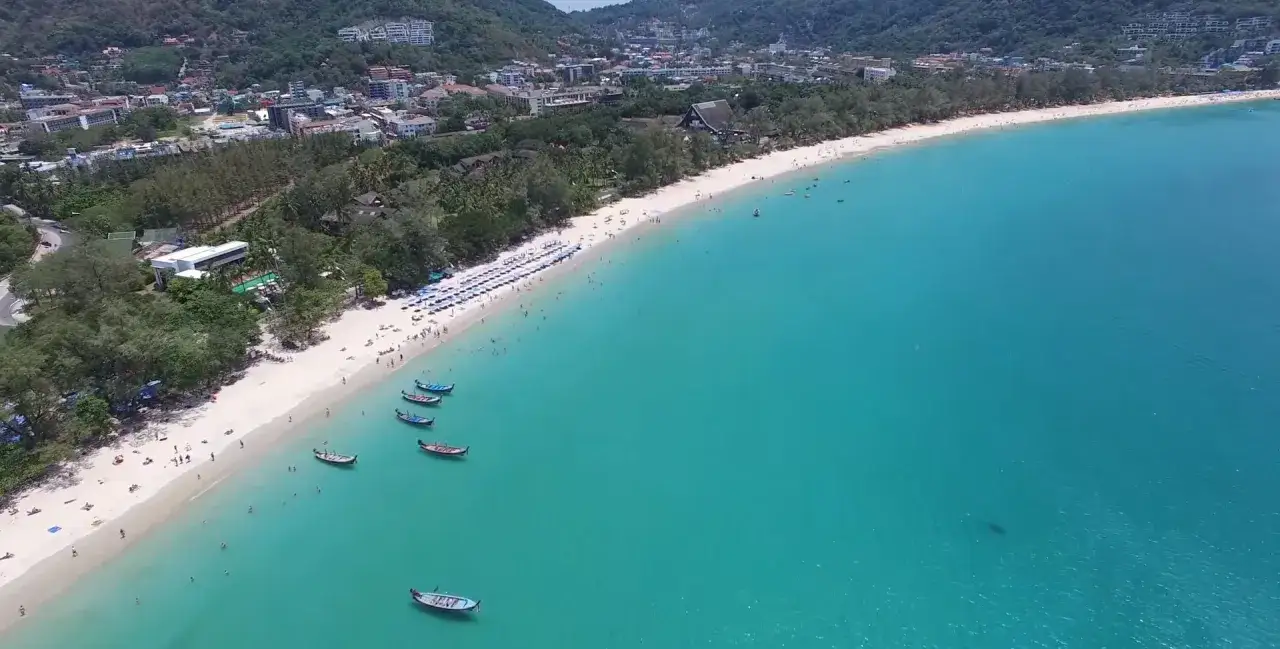 Piękna plaża Kata Beach z białym piaskiem, turkusową wodą i łodziami. W tle widać zielone wzgórza i zabudowania.