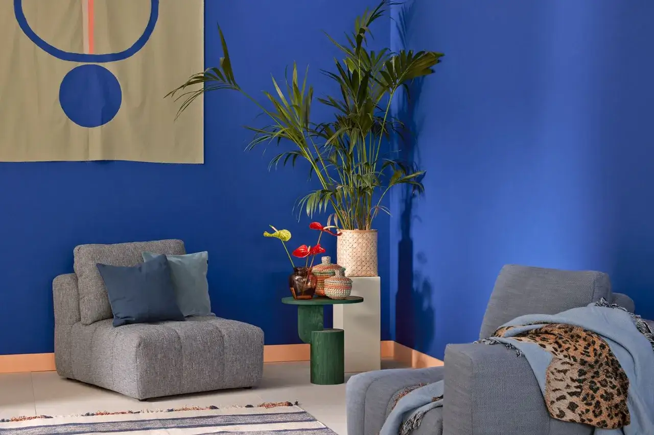 Ein gemütliches Wohnzimmer mit einer leuchtend **blauen Wand**, grauen Sesseln, einer Palme und abstrakter Kunst.