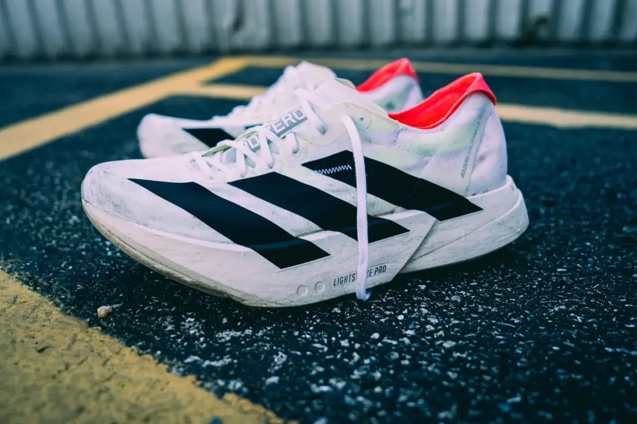 Białe buty do biegania adidas adios pro 4 z czarnymi paskami i neonowym wykończeniem, leżące na asfalcie z żółtymi liniami.