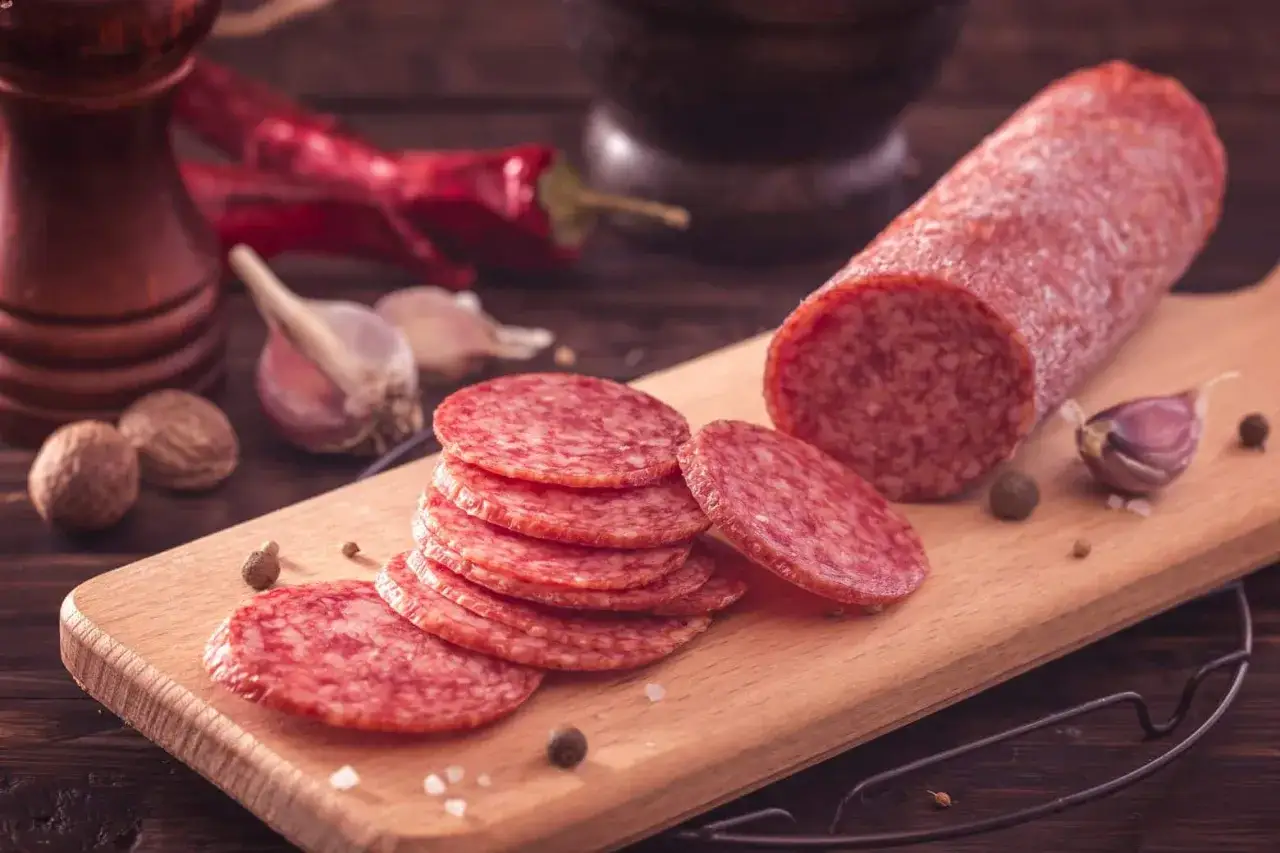 Salami-Scheiben auf Holzbrett, daneben Knoblauch und Chilis. Man kann Salami einfrieren, um sie länger haltbar zu machen.