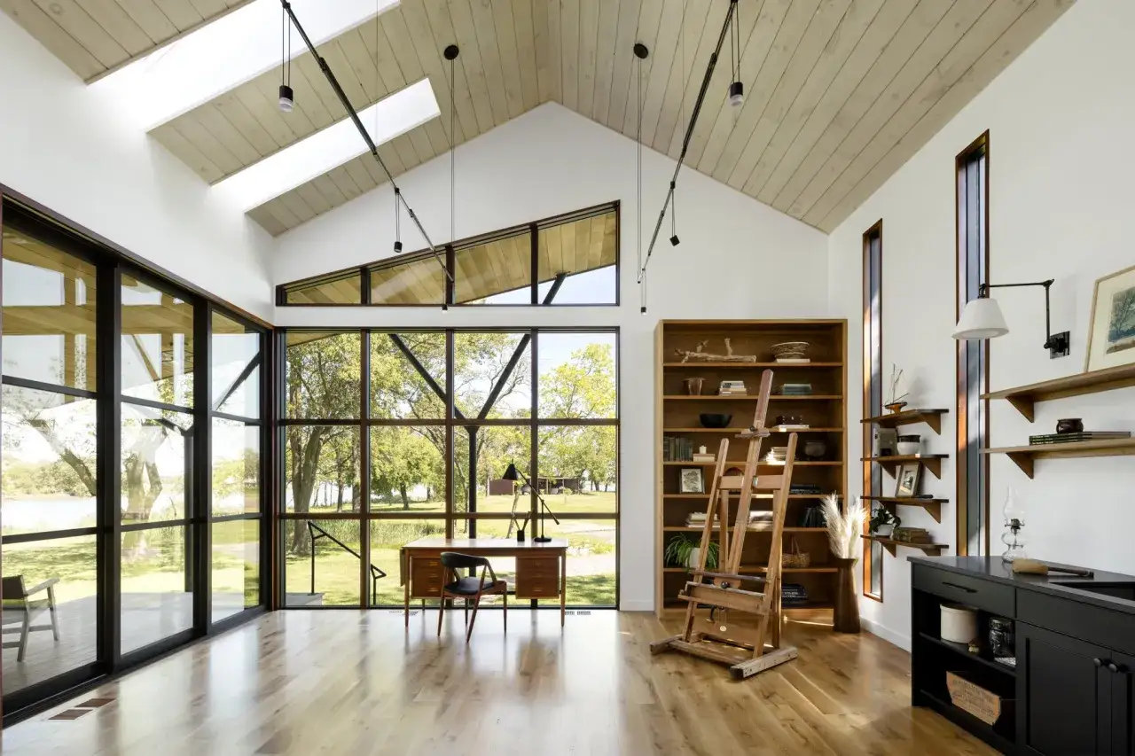 Un inspirador estudio de pintura en casa con grandes ventanales, un caballete de madera y una estanter&iacute;a llena de libros y objetos.
