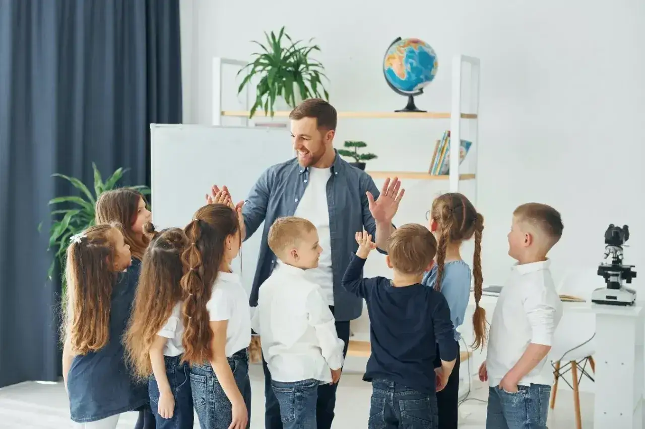 Nauczyciel z dziećmi w klasie, radośnie klaszczą. Czas na wystawienie ocen na 1 semestr!