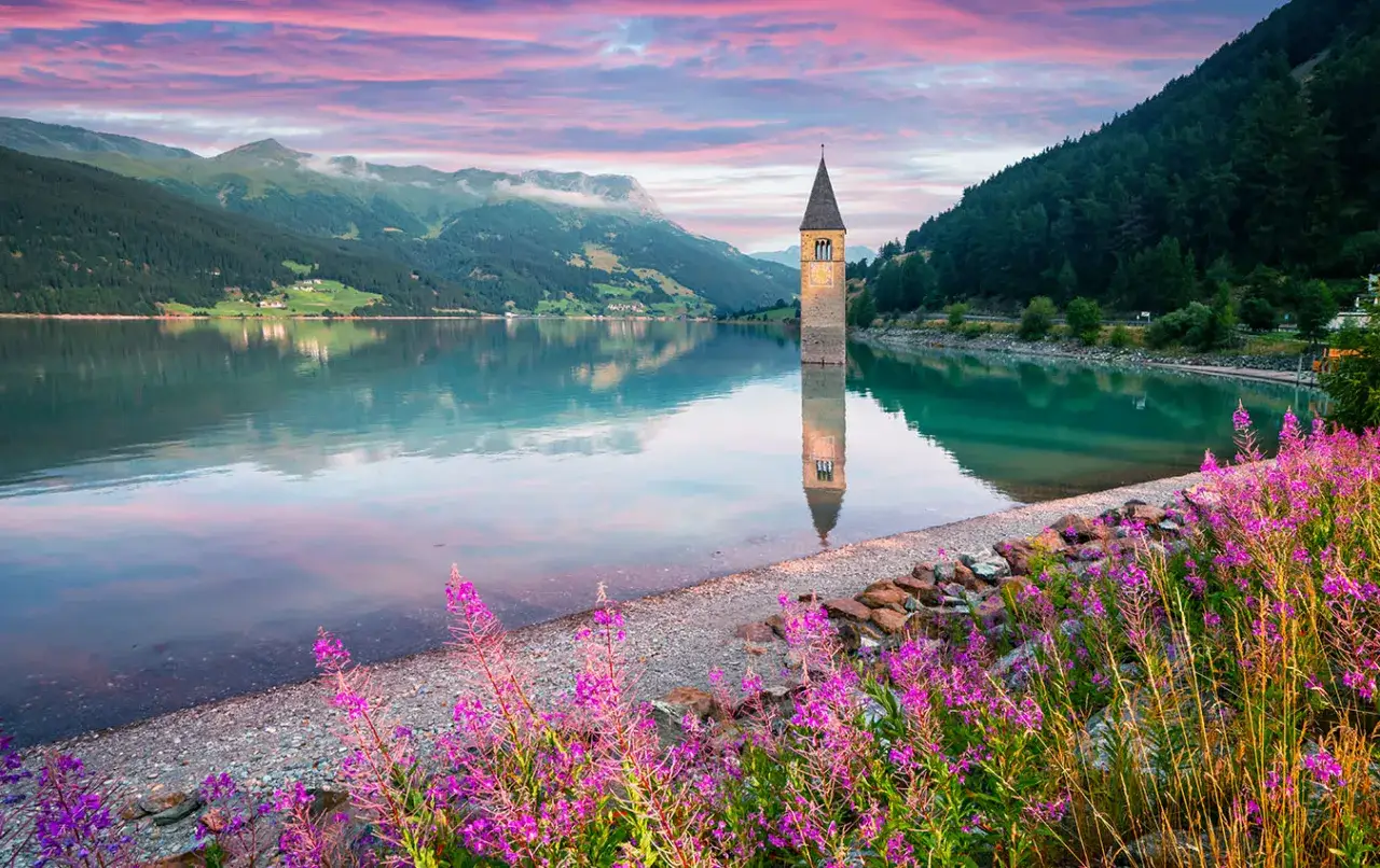 Torre sommersa in un lago alpino, con montagne verdi e cielo rosa all'alba. Un angolo di montagna Friuli Venezia Giulia incantevole.