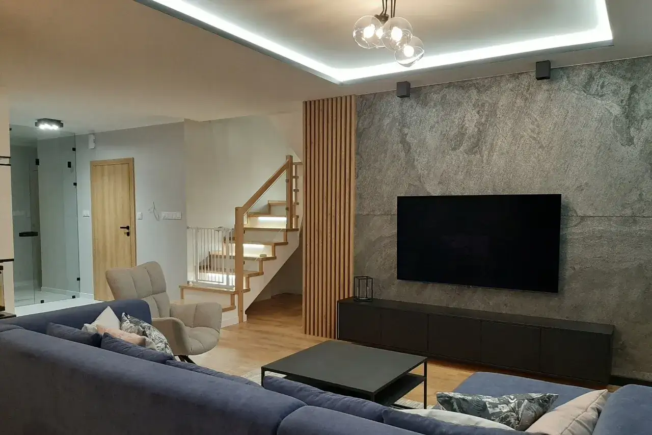 Drewniane lamele tworzą stylową ściankę działową obok schodów. W salonie wygodna kanapa, fotel i telewizor na kamiennej ścianie.