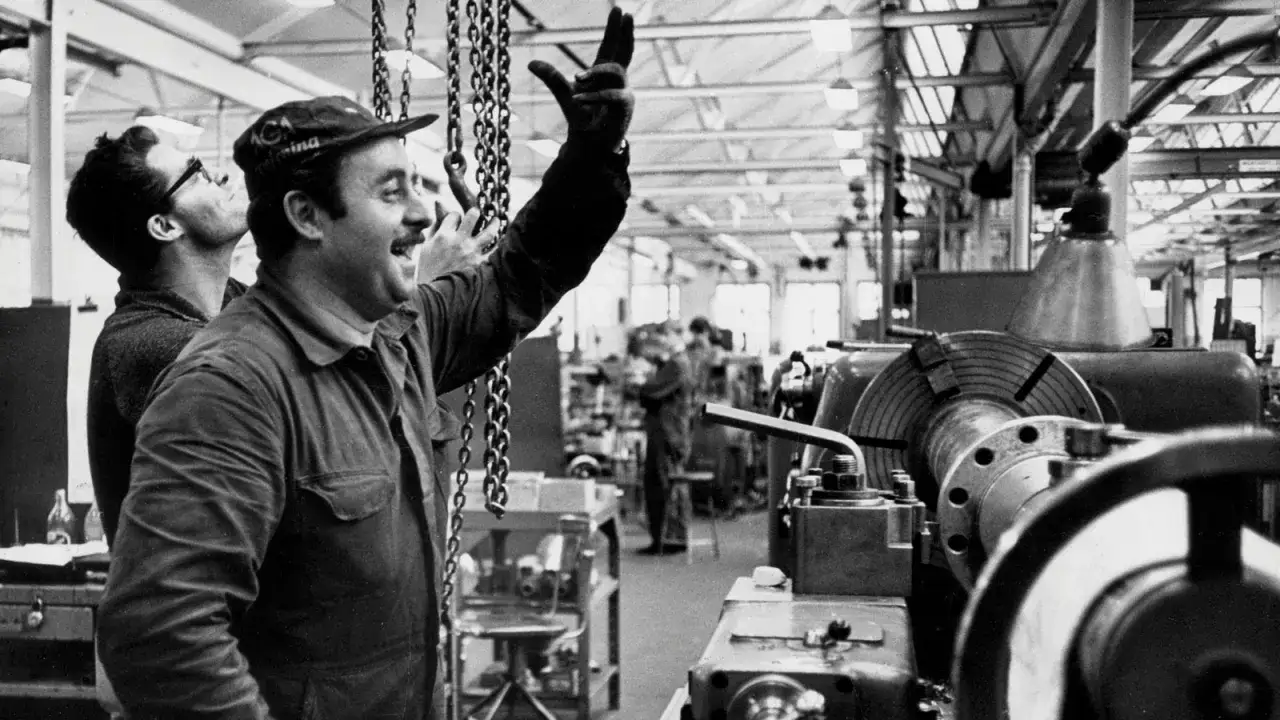 Två män i en verkstad, en gestikulerar glatt. En scen som speglar arbetskraftsinvandring Sverige 1960.