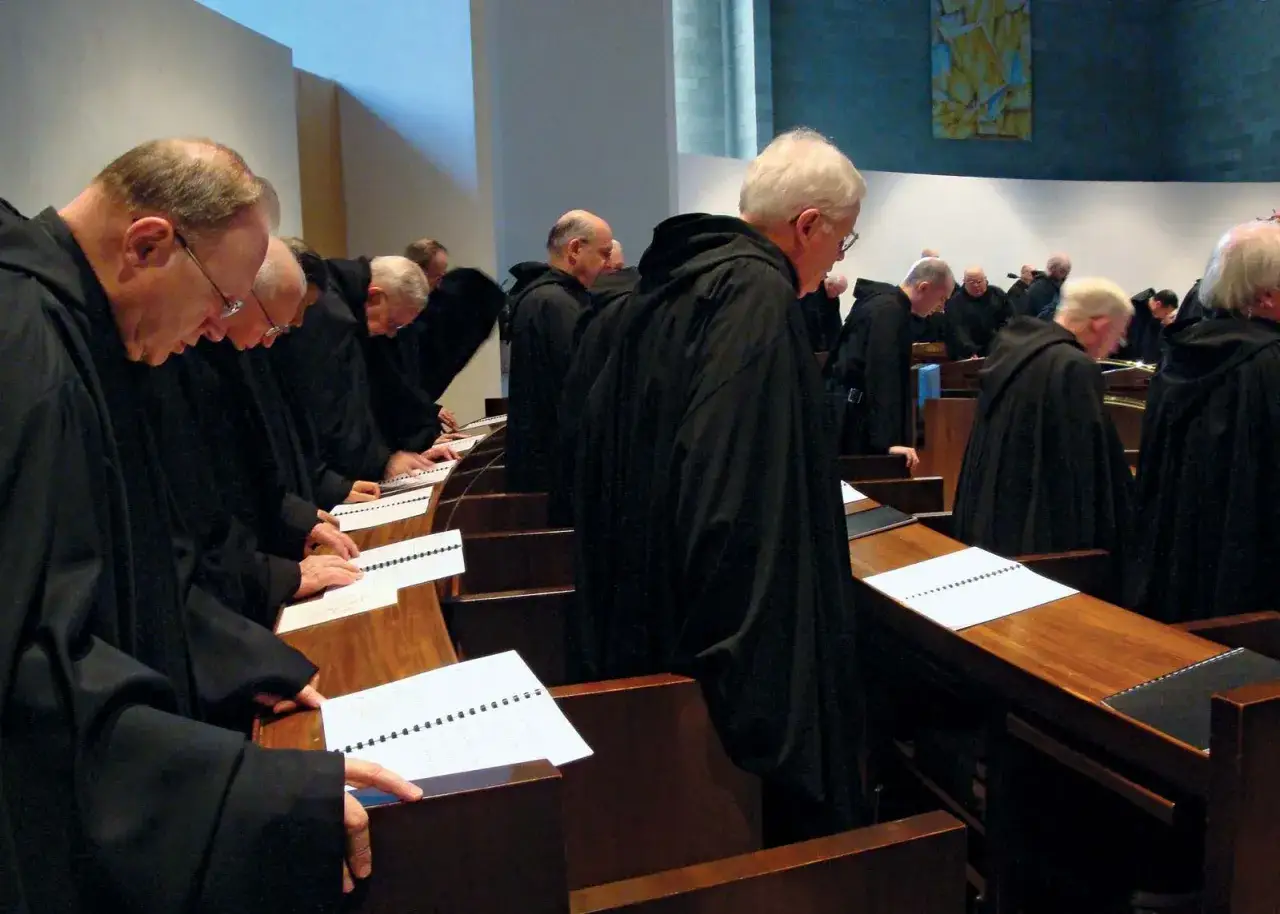 Monks in black robes stand in pews, heads bowed over open books, following the Benedictine calendar.