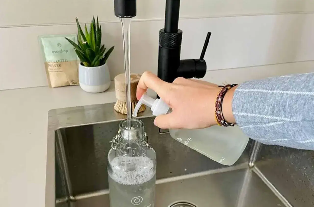 Eine Hand drückt Spülmittel in eine Glasflasche, während Wasser aus dem Wasserhahn läuft. So einfach ist die Trinkflasche reinigen.