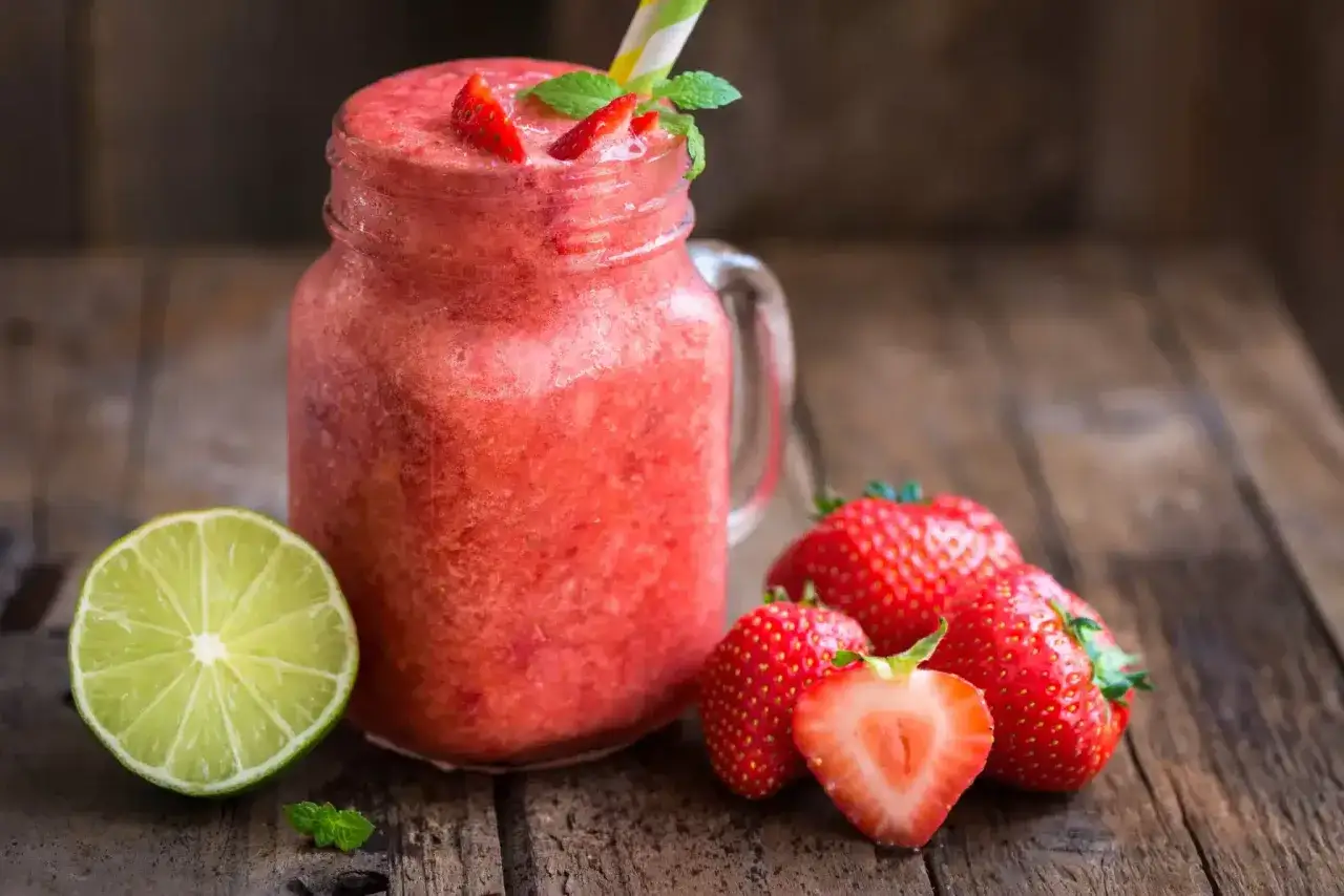 Erfrischendes Erdbeer-Slush-Eis im Glas, garniert mit frischen Erdbeeren und Minze, daneben eine Limettenspalte und weitere Erdbeeren.