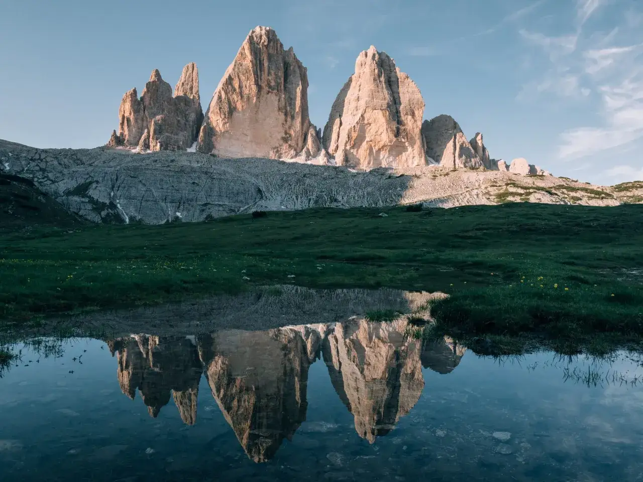 Trzy majestatyczne szczyty Tre Cime di Lavaredo odbijają się w spokojnej tafli wody, otoczone zieloną łąką.