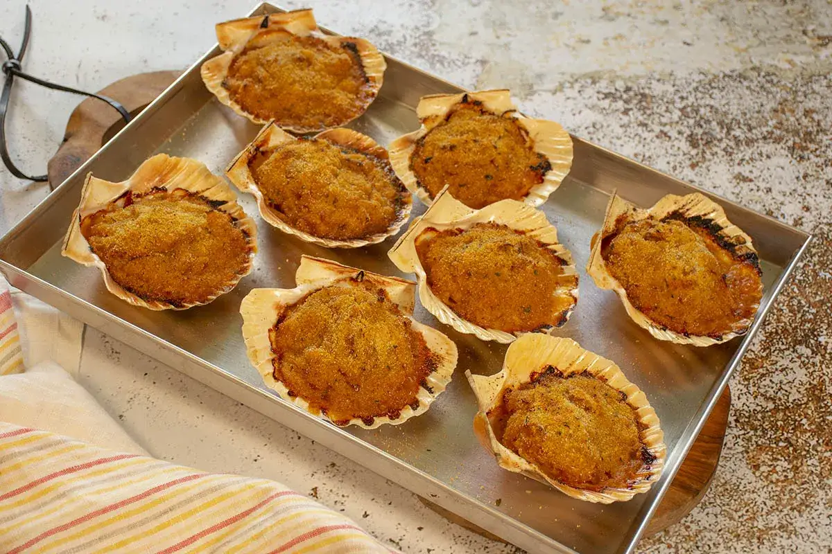 Ocho vieiras gratinadas, doradas y crujientes, servidas en sus conchas sobre una bandeja metálica.