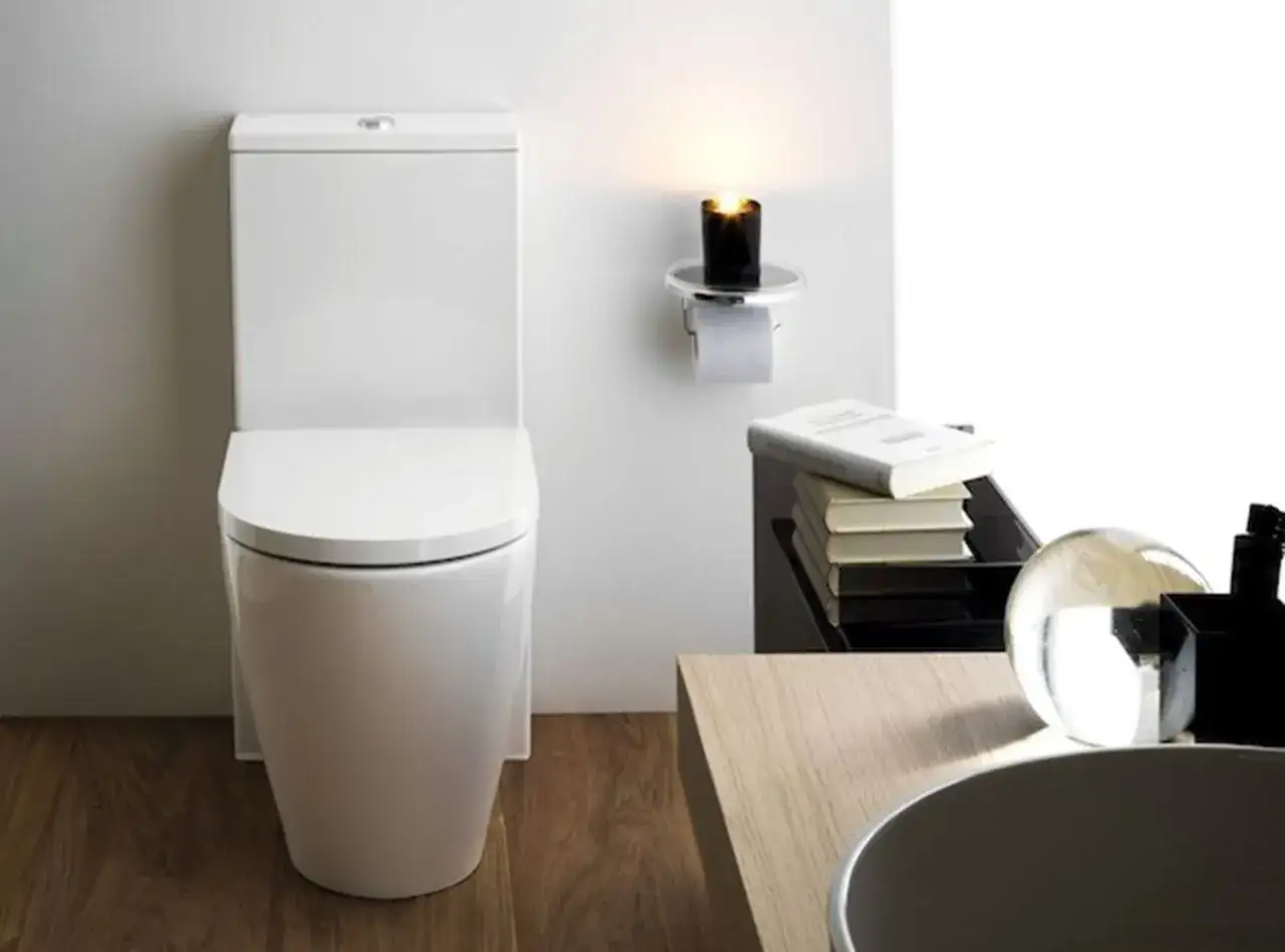 Elegancki, biały wc kompakt nowoczesny. Obok świeca i stos książek, tworząc przytulną atmosferę.