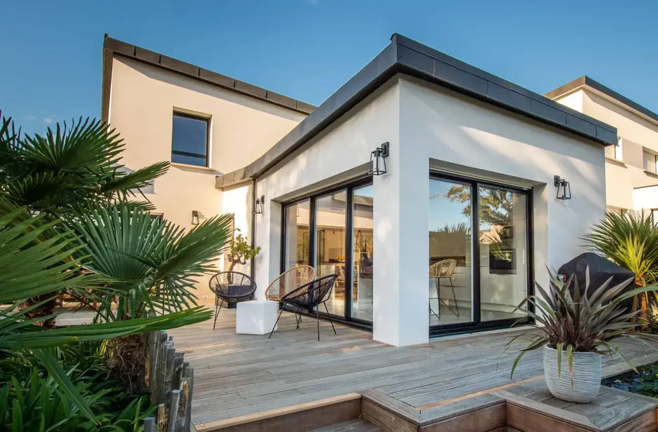 Belle extension maison avec baie vitrée donnant sur une terrasse en bois. Palmiers et mobilier d'extérieur invitent à la détente.