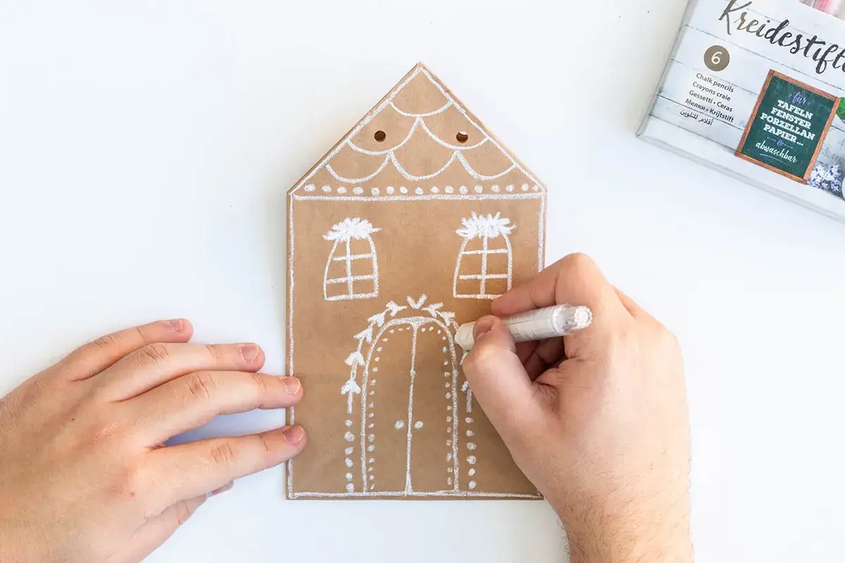 Mit weißen Kreidestiften werden weihnachts giveaways selbstgemacht. Ein Haus aus braunem Papier wird verziert.