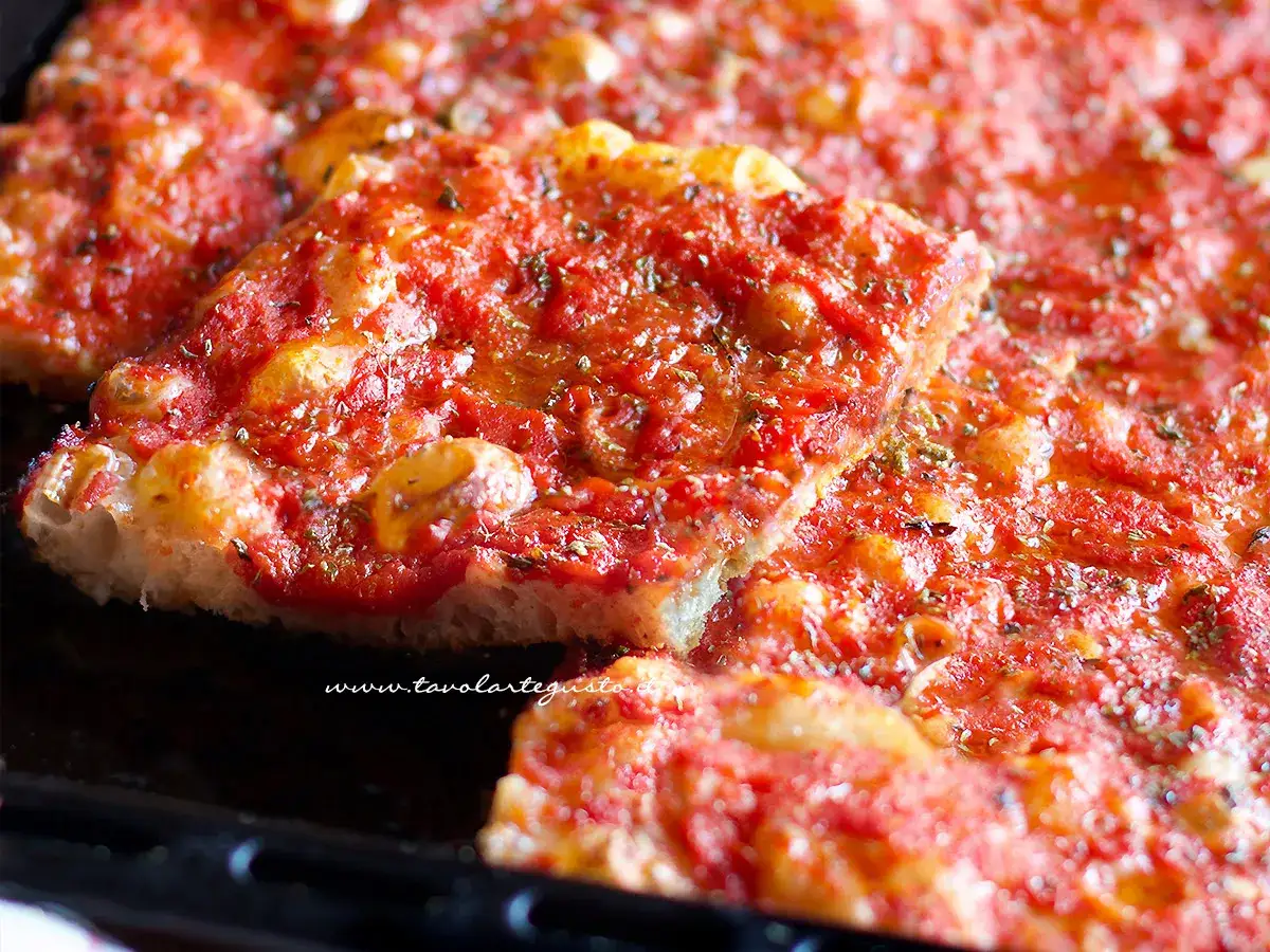 Una fetta di pizza in teglia, con pomodoro e formaggio filante, cosparsa di erbe aromatiche.