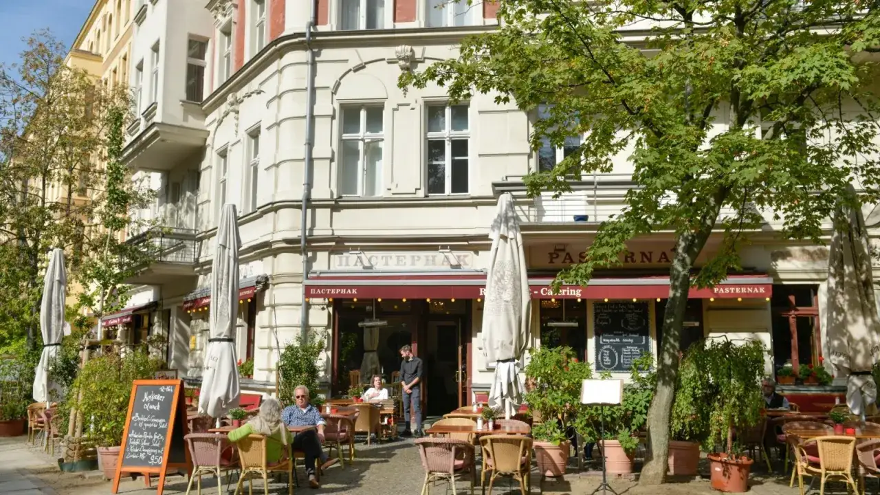 Gem&uuml;tliches **Prenzlauer Berg Restaurant** mit Au&szlig;enbereich. G&auml;ste sitzen an Tischen unter B&auml;umen, die Markise ist rot.