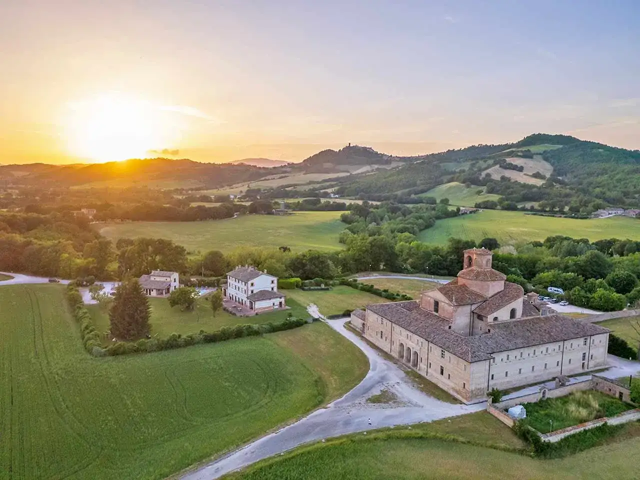Un tramonto dorato illumina dolci colline e un complesso monastico a Frontino, Marche. Campi verdi e alberi circondano antichi edifici.