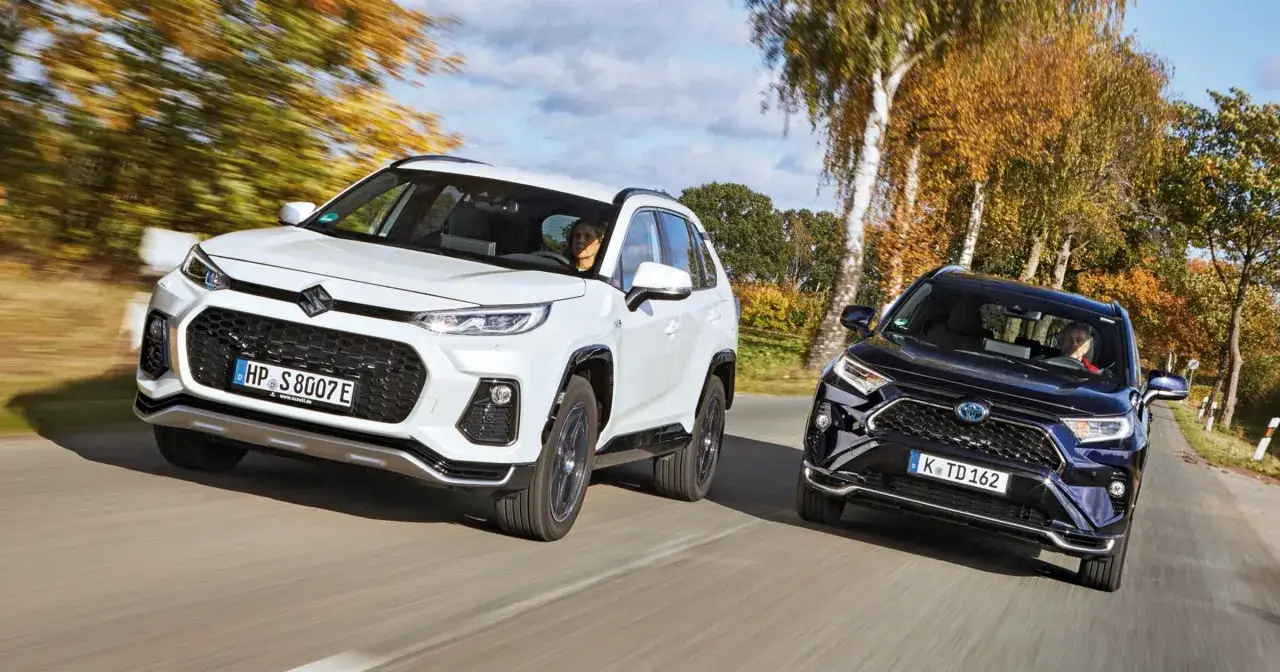 Biały i ciemny SUV jadą obok siebie na drodze, otoczone jesiennymi drzewami. Wyglądają jak modele z przyszłości, być może zapowiedź **rav4 2026**.