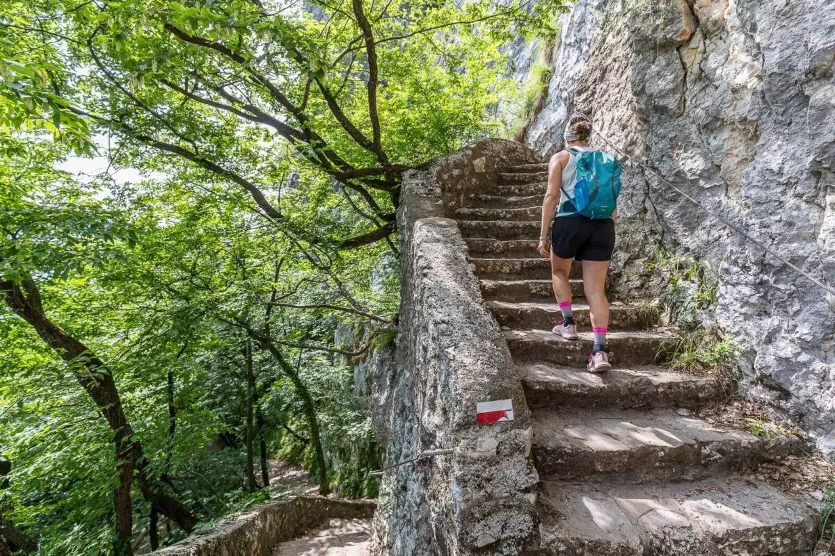 Una donna con zaino blu sale una scalinata di pietra, parte del sentiero della speranza, tra alberi verdi e roccia.