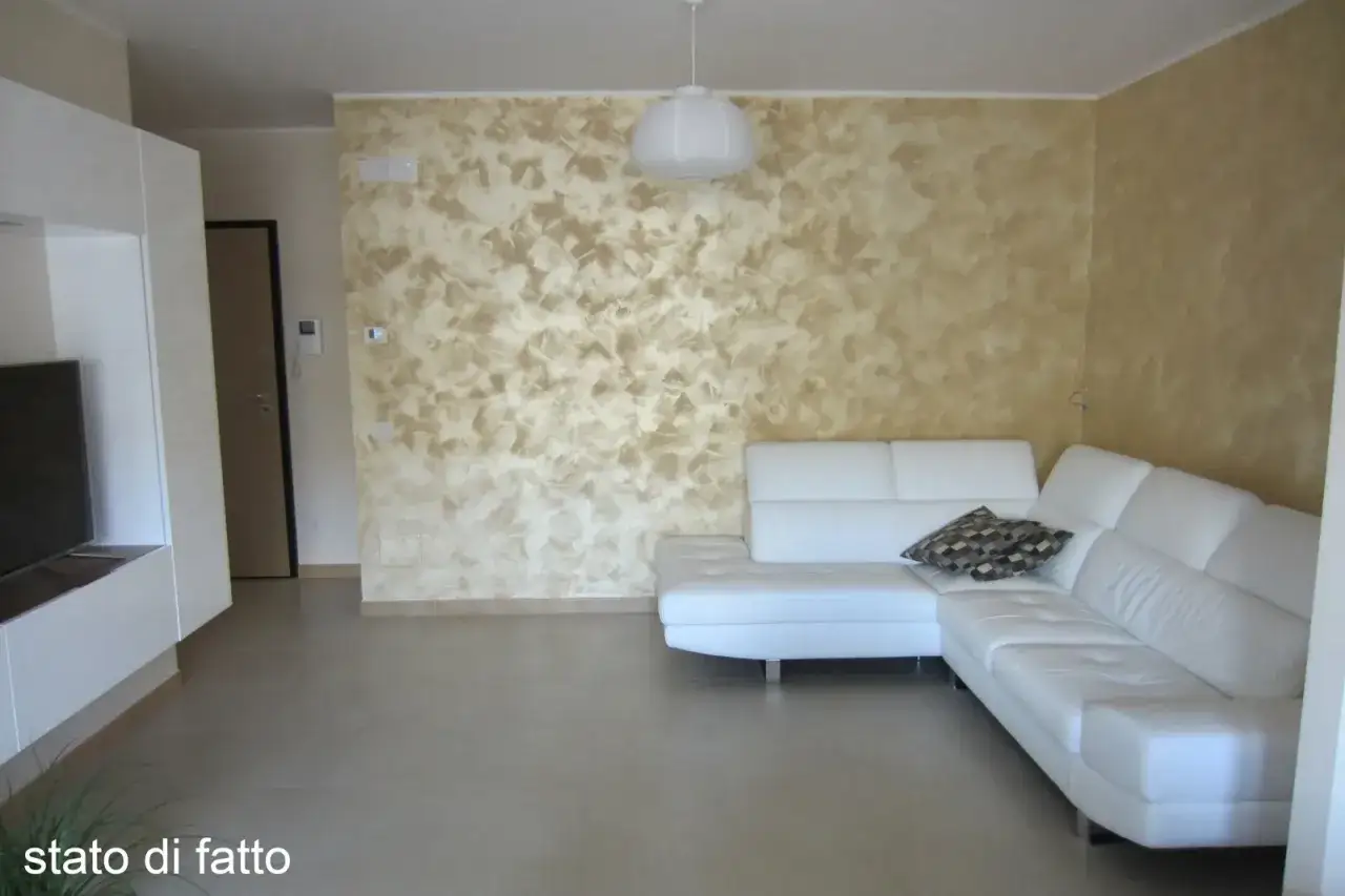 Soggiorno moderno con divano bianco angolare e parete decorata con un effetto materico che crea una tavola abbinamento colori calda e accogliente.