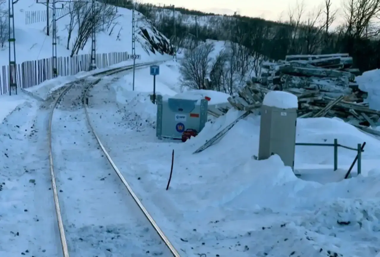 Snöiga järnvägsspår leder mot en skogsklädd kulle. En hög med timmer ligger vid sidan av spåret, redo för ett tåg till Luleå eller Kiruna.