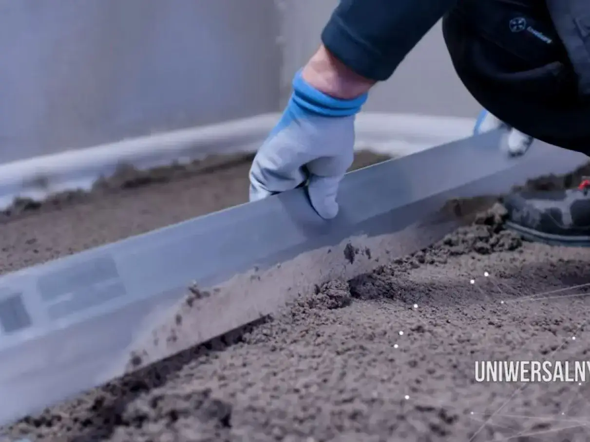 Ręka w rękawicy wyrównuje podłoże za pomocą łaty. Szybki beton gotowy do dalszych prac.
