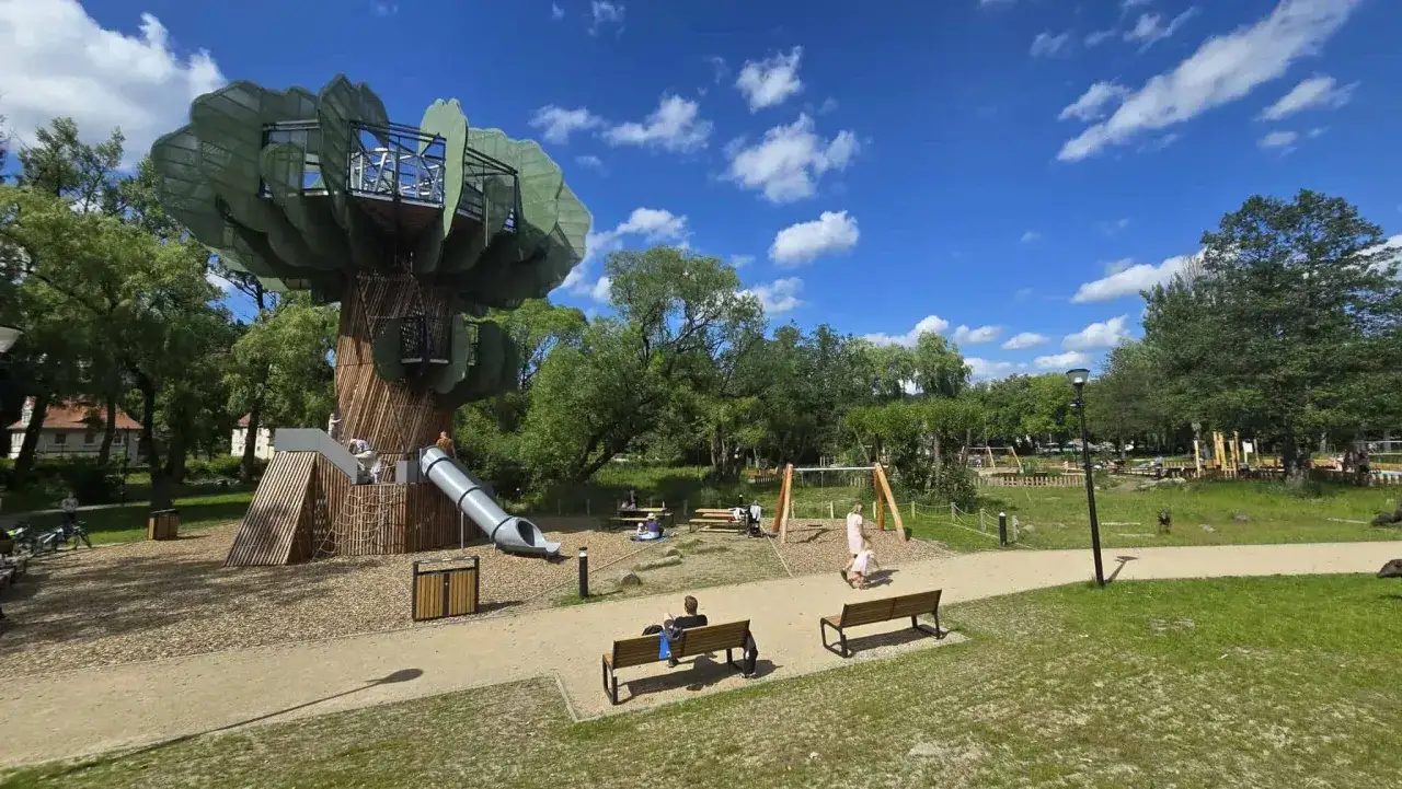 Wielka, bajkowa konstrukcja przypominająca drzewo z domkami i zjeżdżalnią to główne duszniki zdrój atrakcje dla dzieci. W tle huśtawki i zielony park.