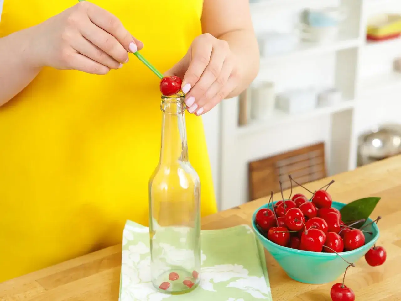 Hände entkernen Kirschen mit einem grünen Strohhalm und stecken sie in eine Glasflasche. Daneben eine Schüssel voller Kirschen.
