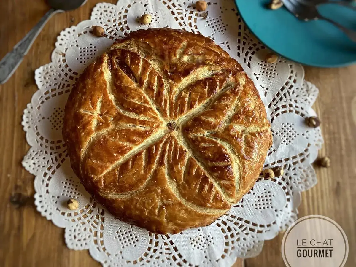 Galette frangipane noisette dor&eacute;e, d&eacute;cor&eacute;e d'une feuille stylis&eacute;e, sur une nappe en dentelle. Quelques noisettes enti&egrave;res pars&egrave;ment la table.