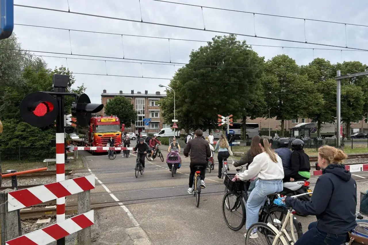 Fietsers wachten bij een spoorwegovergang met rood licht. Een vrachtwagen staat stil, terwijl fietsers geduldig wachten om door rood te rijden.
