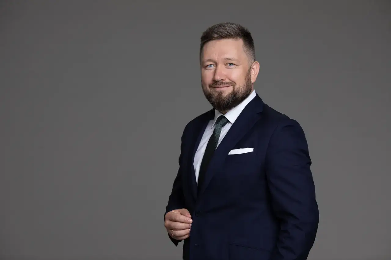 Mężczyzna w granatowym garniturze i zielonym krawacie, uśmiecha się do kamery.