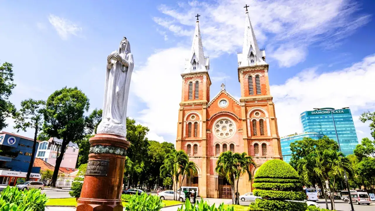 Katedra Notre Dame w Ho Chi Minh, z posągiem Matki Boskiej na pierwszym planie, w otoczeniu zieleni i nowoczesnych budynk&oacute;w.