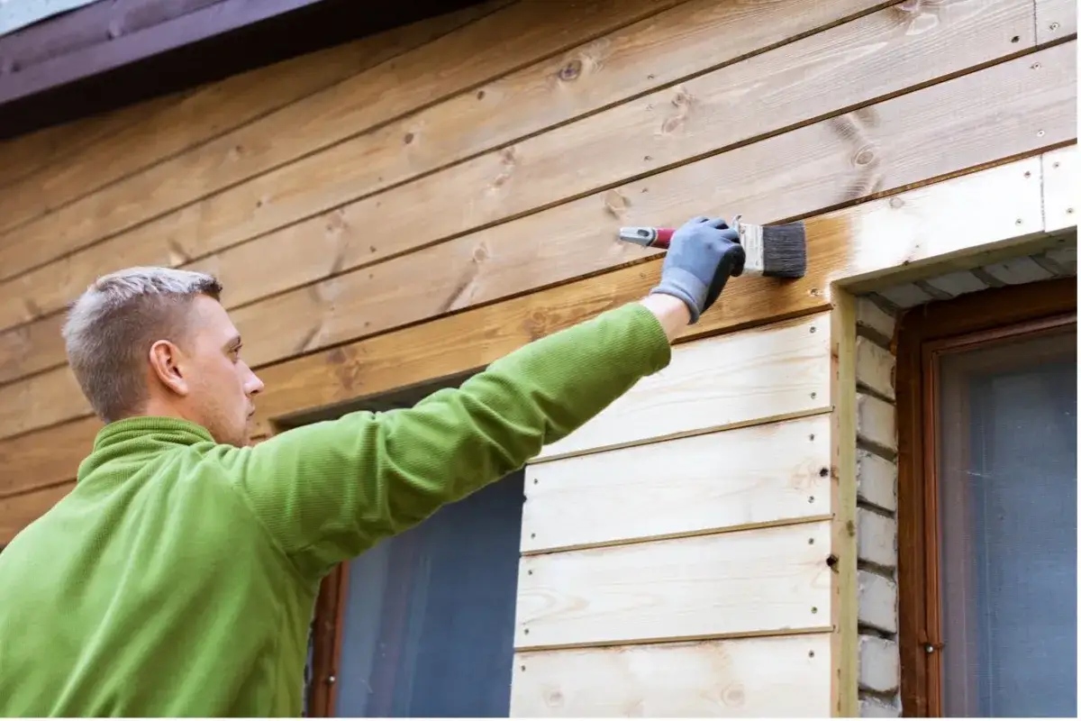 Man målar plank med pensel. Träfasaden får ny färg och skydd.