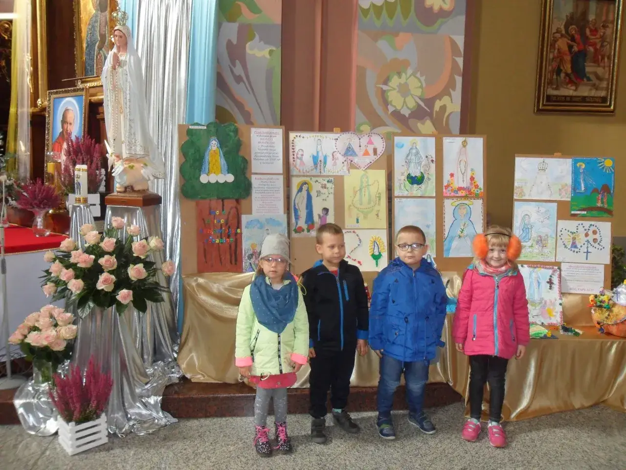 Dzieci stoją przed wystawą prac plastycznych z Matką Bożą. Chłopcy i dziewczynka w kurtkach, jakby chcieli powiedzieć: