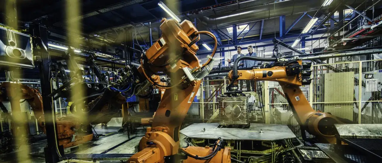 Pomarańczowe ramiona robotów pracują w fabryce, demonstrując zaawansowaną mechatronikę. Dwóch inżynierów obserwuje proces.