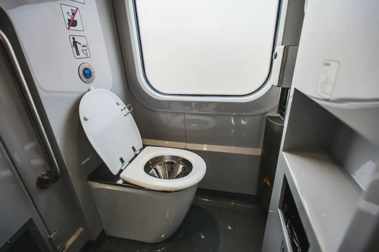 Wnętrze toalety w pociągu Pendolino. Biała muszla klozetowa z otwartą klapą, obok okno i uchwyt.
