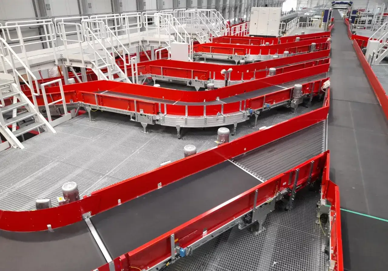 System taśmociągów w centrum logistycznym. Czerwone taśmy transportowe wiją się, tworząc skomplikowany labirynt, gotowy do obsługi przesyłek.