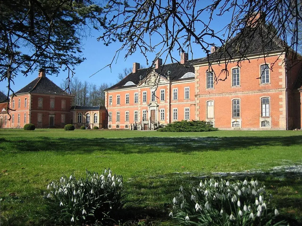 Schloss Bothmer, ein prächtiges Backsteingebäude, erhebt sich vor einem klaren blauen Himmel. Schneeglöckchen blühen im Vordergrund.