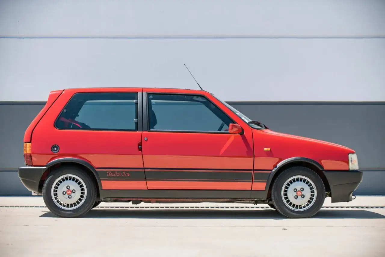 Czerwony Fiat Uno Turbo i.e. z czarnymi pasami na boku, zaparkowany na tle szarej ściany.