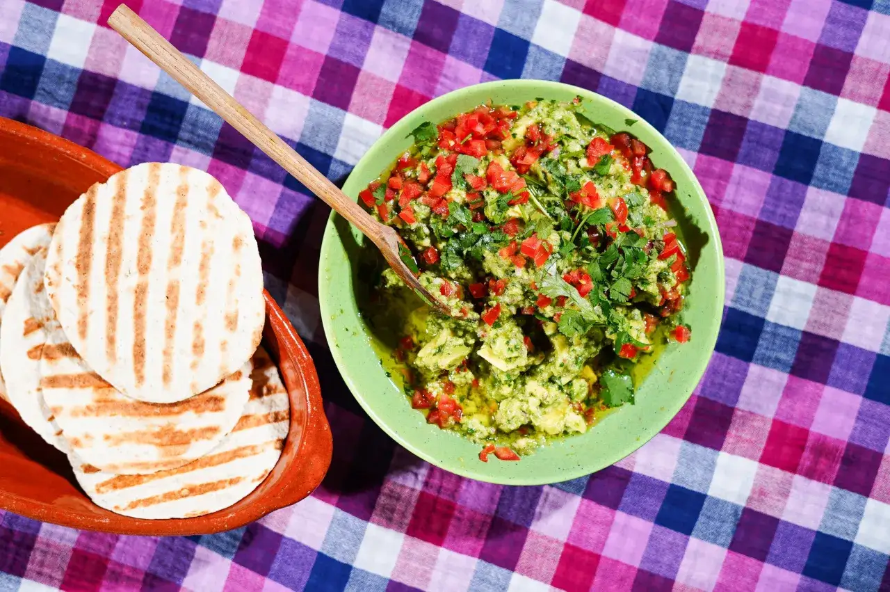 Un bol de recette guacamole traditionnel, garni de dés de tomates et de coriandre fraîche, à côté de tortillas grillées dans un plat en terre cuite.