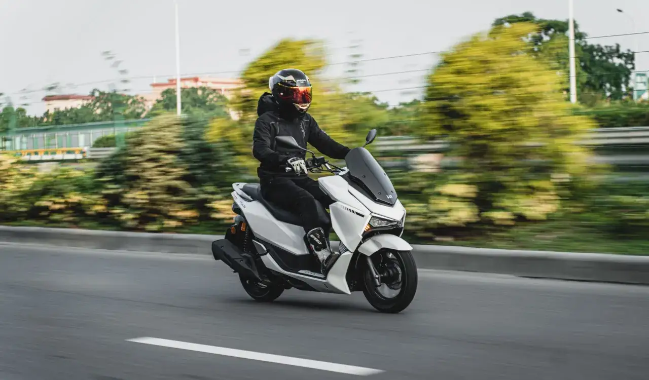 Persona con carnet B moto conduce un scooter blanco por una carretera con vegetaci&oacute;n borrosa.