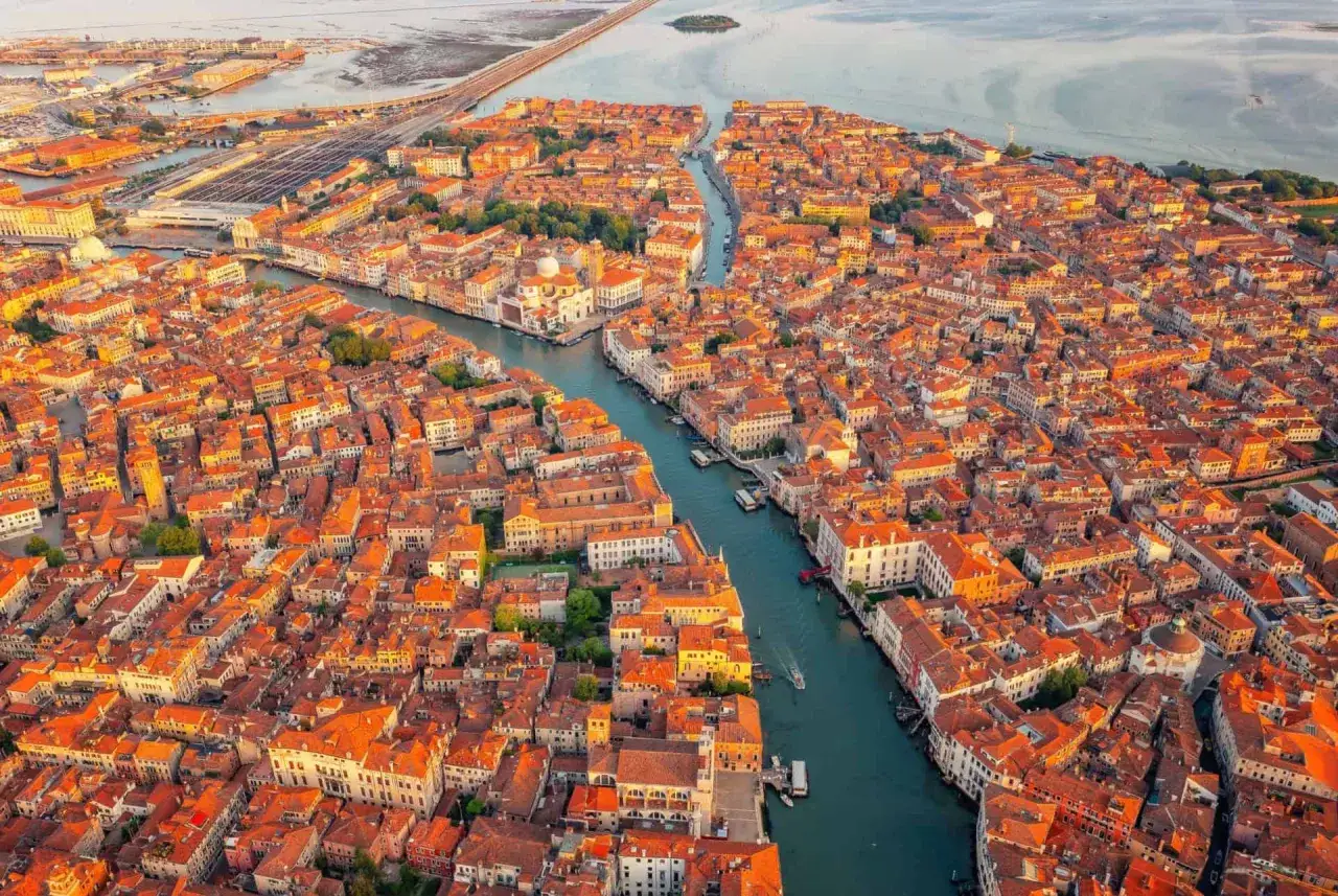 Vue a&eacute;rienne de Venise au coucher du soleil, avec ses canaux sinueux et ses toits orang&eacute;s. Un pont embl&eacute;matique de Venise se dessine au loin.