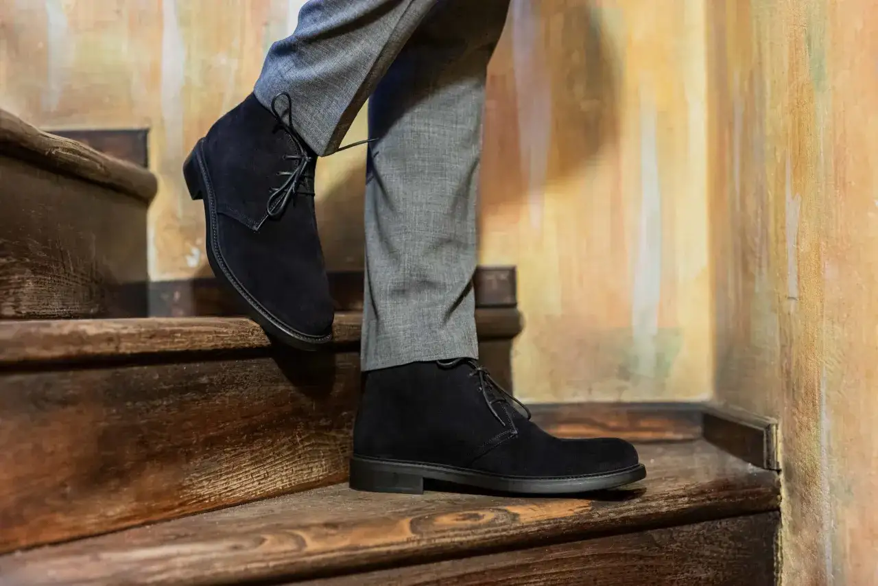 Un uomo indossa eleganti polacchi scamosciati neri e pantaloni grigi, mentre scende una scala di legno.