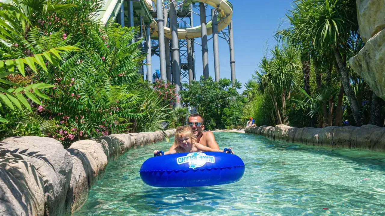 Un père et sa fille profitent d'une descente en bouée dans la rivière d'un centre de vacances et de loisirs, entourés de végétation luxuriante et de toboggans aquatiques.