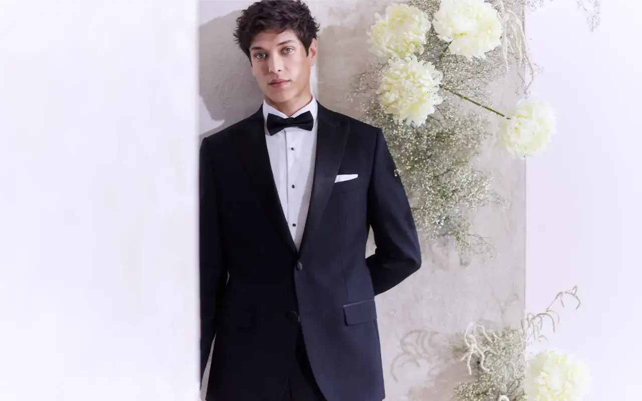 A young man in a tuxedo, perfect for black tie weddings, poses with white flowers.