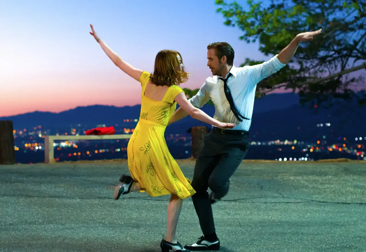 Para tańczy w żółtej sukience i białej koszuli na tle miasta nocą, jakby w magicznym La La Land.