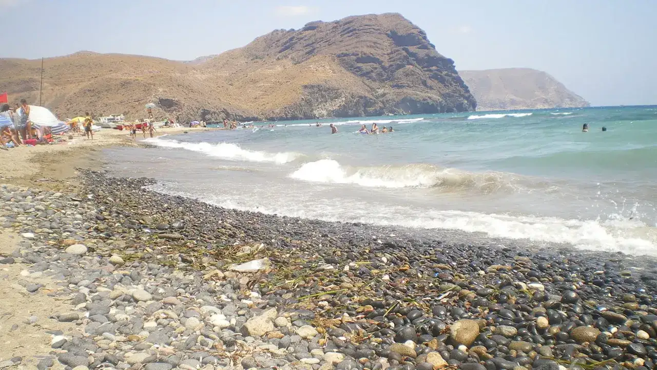 Gente disfrutando de un día soleado en las playas de **las negras cabo de gata**. Olas rompen en la orilla pedregosa, con montañas áridas de fondo.