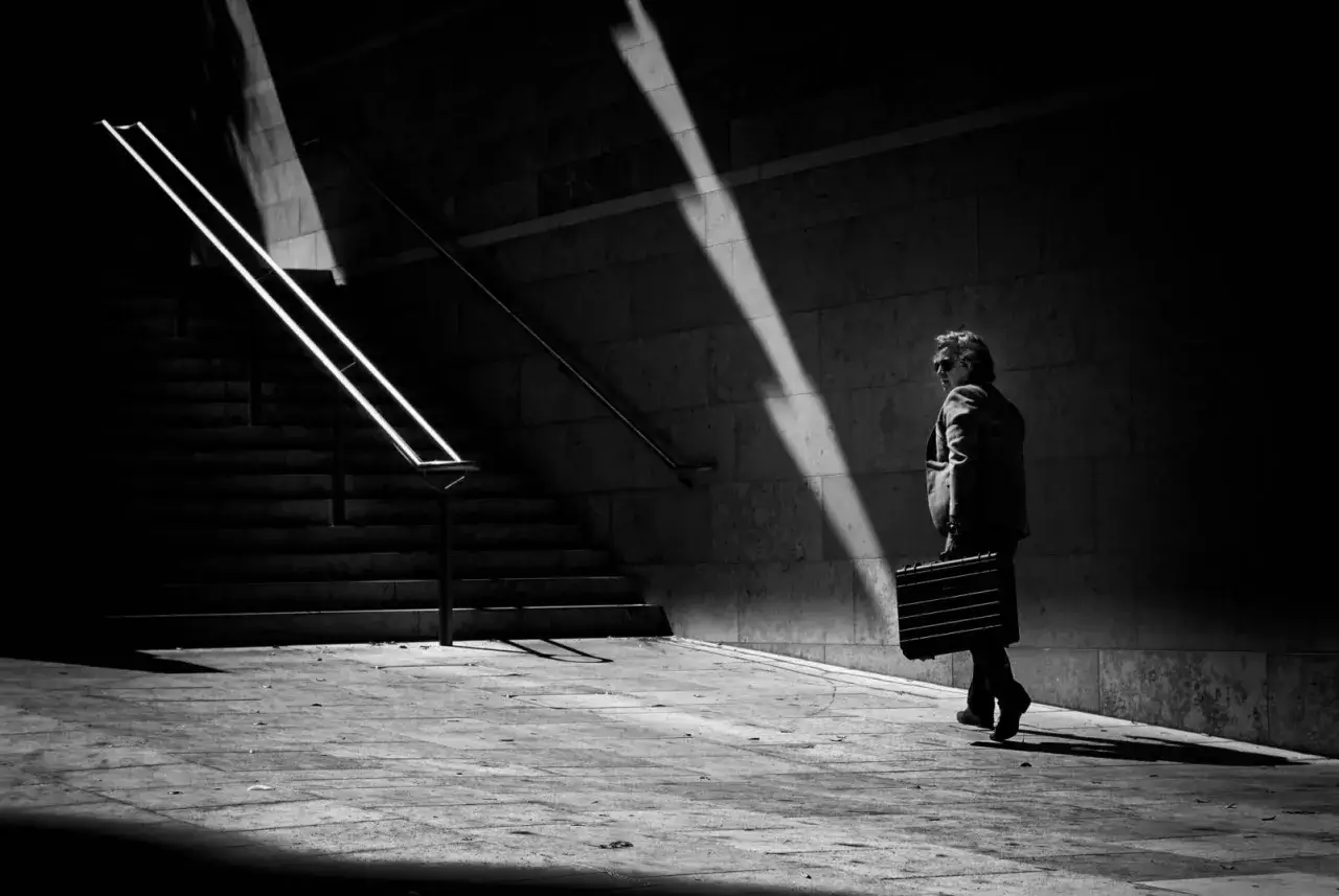 A man in a suit carries a briefcase, walking away from illuminated stairs in a stark, high-contrast scene, reminiscent of Trent Parke's urban narratives.